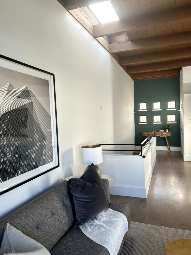Modern living space with gray couch, abstract wall art, lamp, wooden ceiling, skylight, and a hallway with small square windows and a console table.