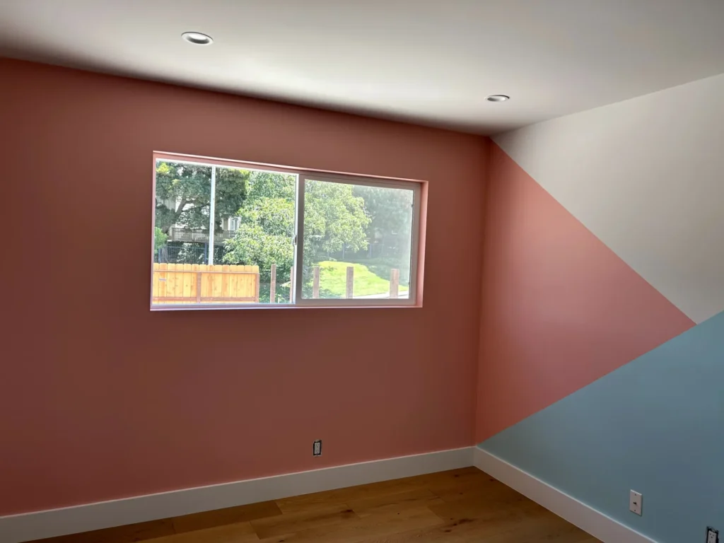 A room with pink, blue, and white geometric painted walls, a wooden floor, and a window showing trees and a fence outside.
