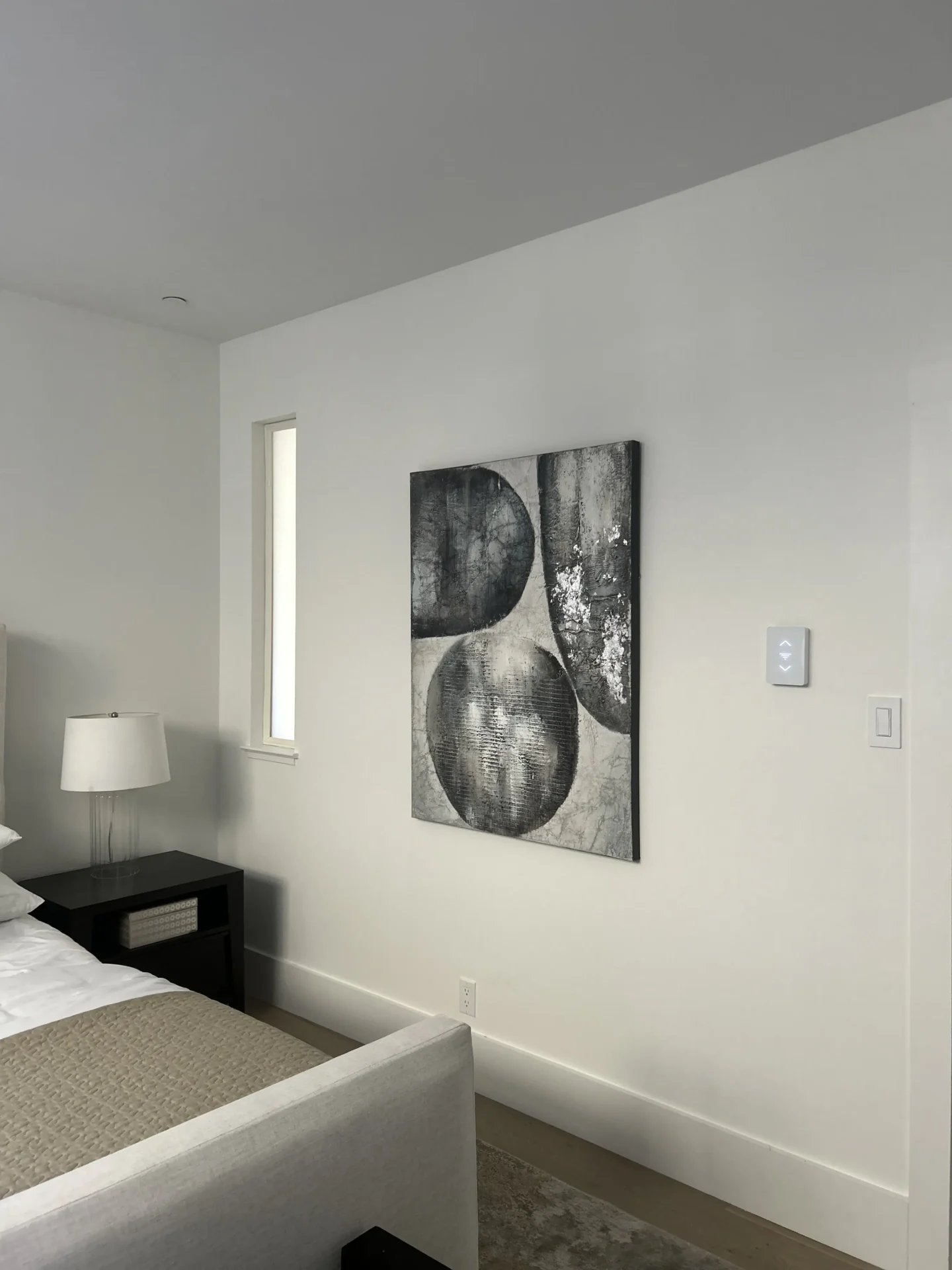 A modern bedroom with a bed, nightstand, lamp, and abstract black and gray artwork hanging on a white wall.