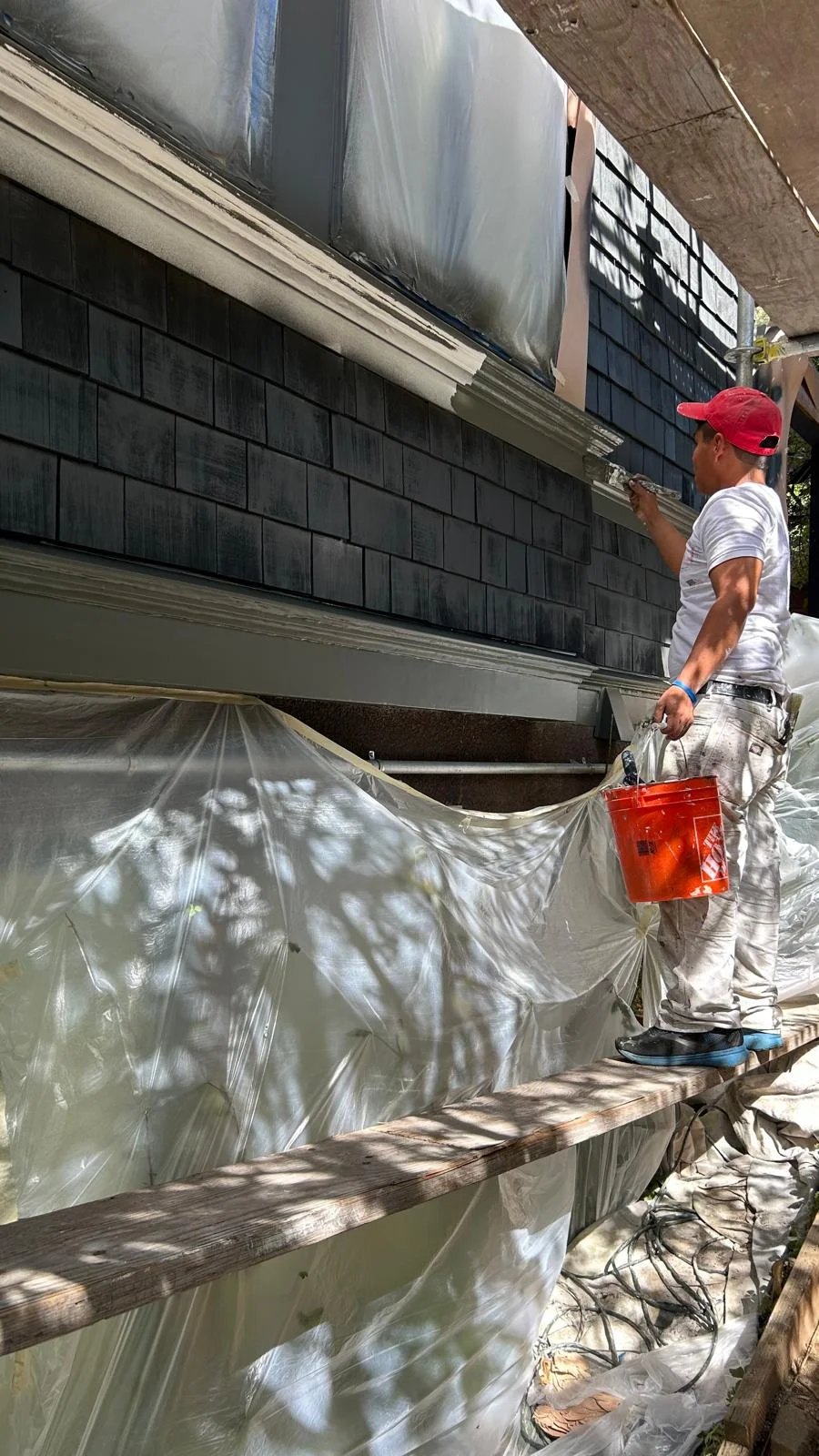 A person standing on scaffolding paints dark gray shingles on a house exterior, with plastic sheets covering areas below for protection, highlighting painters Berkeley CA in action.