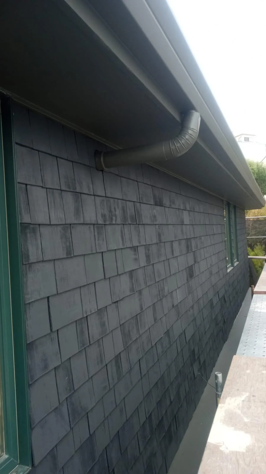 A building exterior with dark slate tiles covering the wall and a black pipe protruding from under the roof gutter, serviced by a painting contractor Mill Valley.