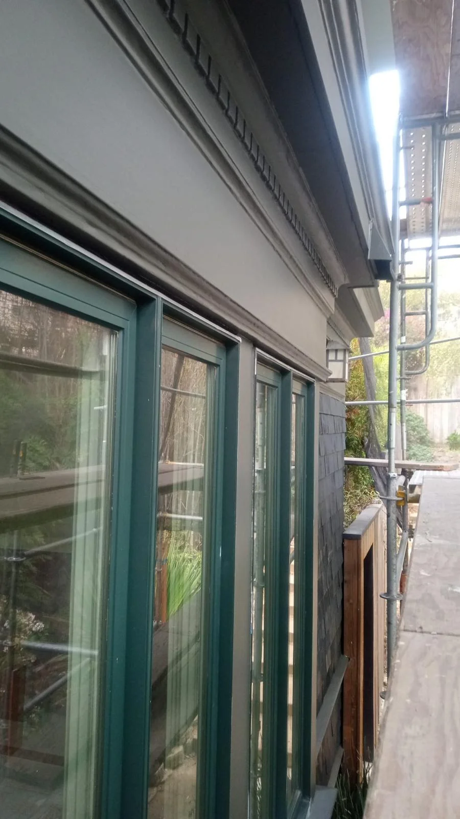 Green-framed windows beneath detailed exterior trim, with scaffolding and a walkway beside a wooden fence and stone wall, showcasing painters San Francisco at work.
