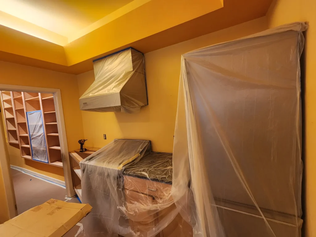 A kitchen under renovation with appliances and cabinetry covered in plastic sheeting painting services san francisco and a drill on the countertop.